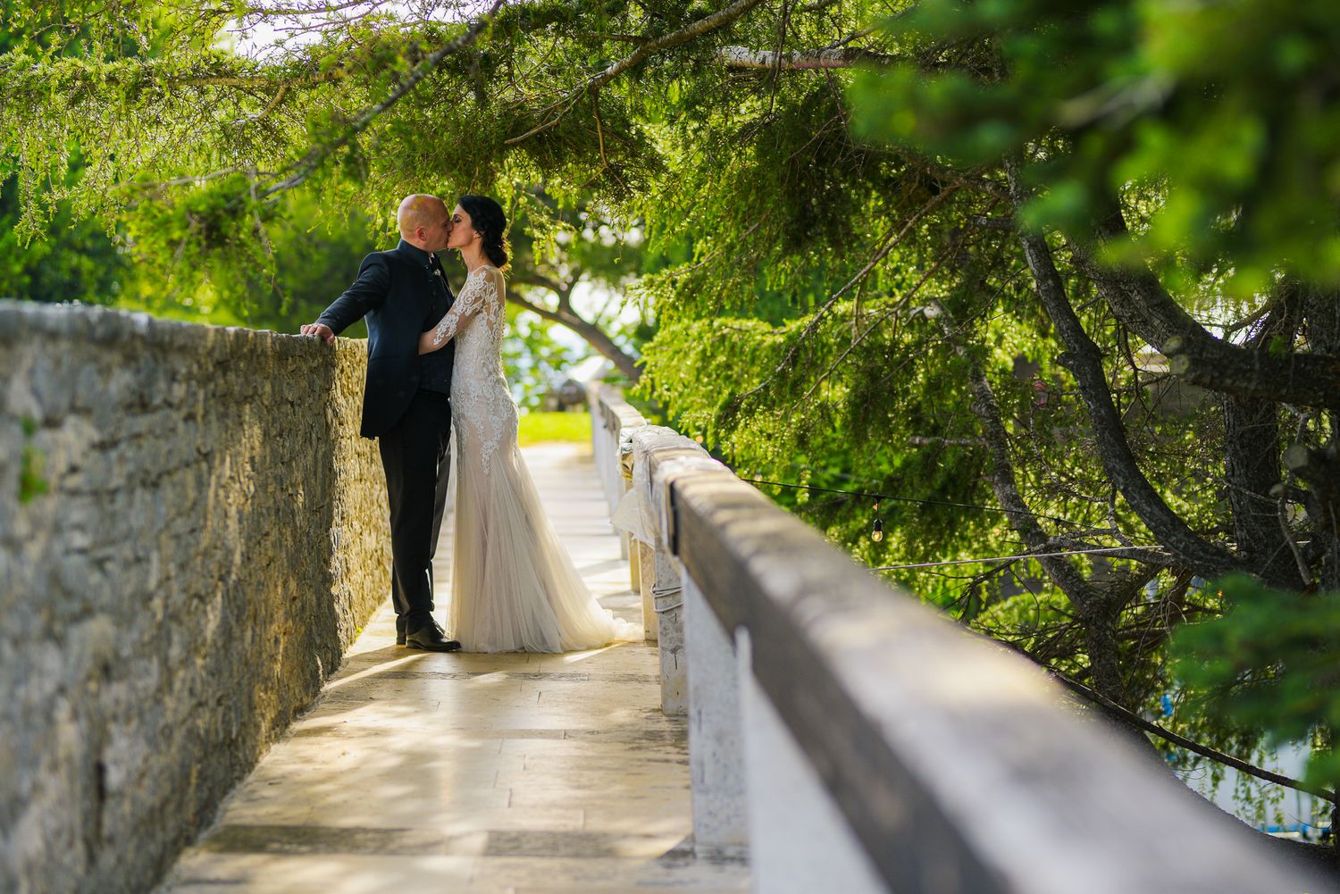 Bride and groom share a romantic moment on a stone bridge surrounded by lush green trees.