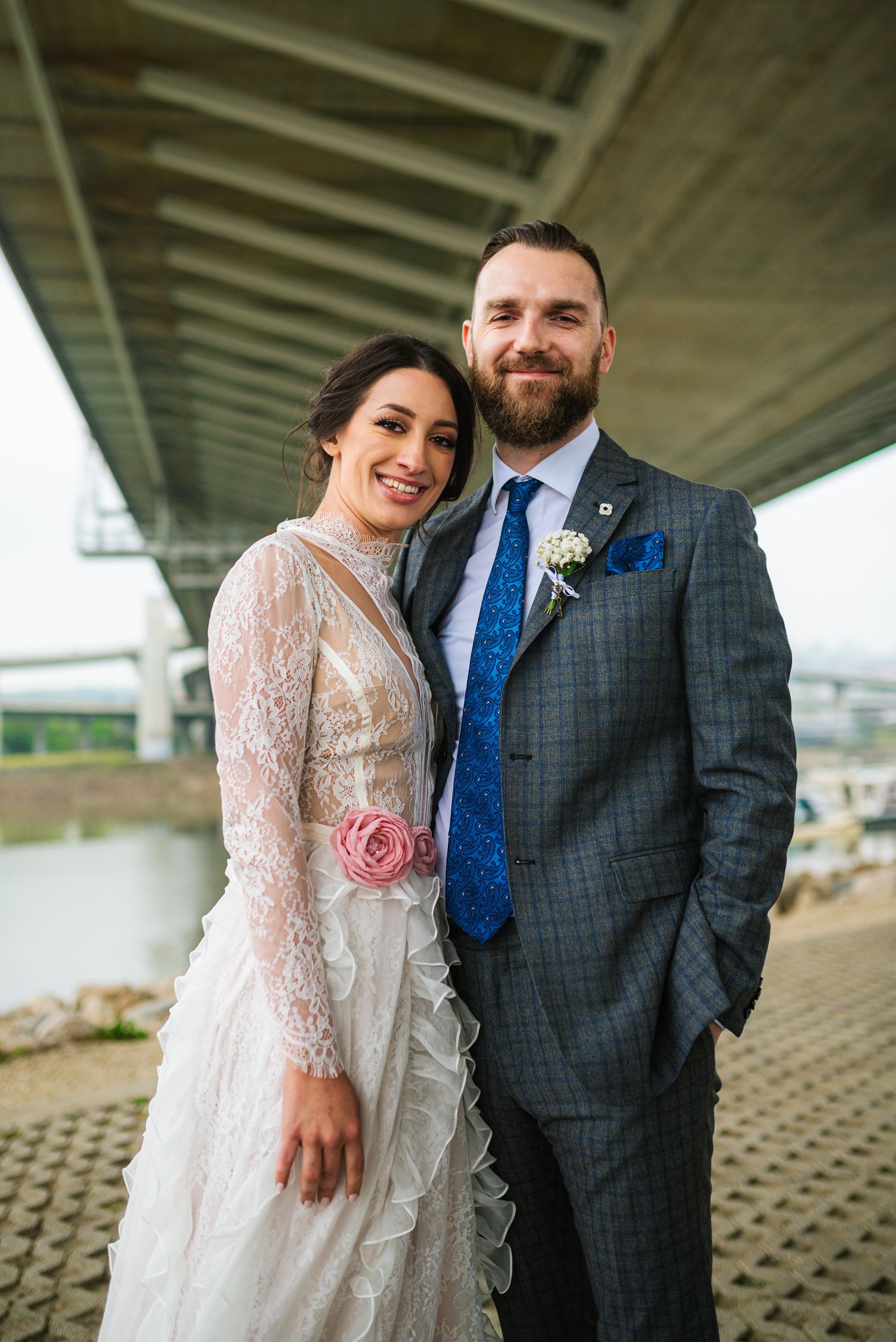 Smiling bride in lace dress and bearded groom in gray suit posing together under bridge on wedding day.