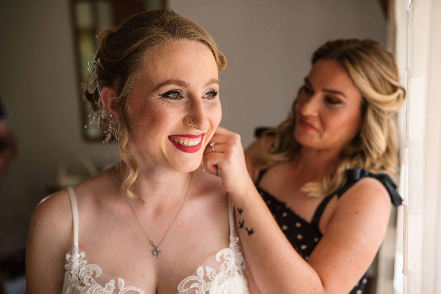 Bridesmaid helping bride put on earrings while she smiles in lace wedding dress.