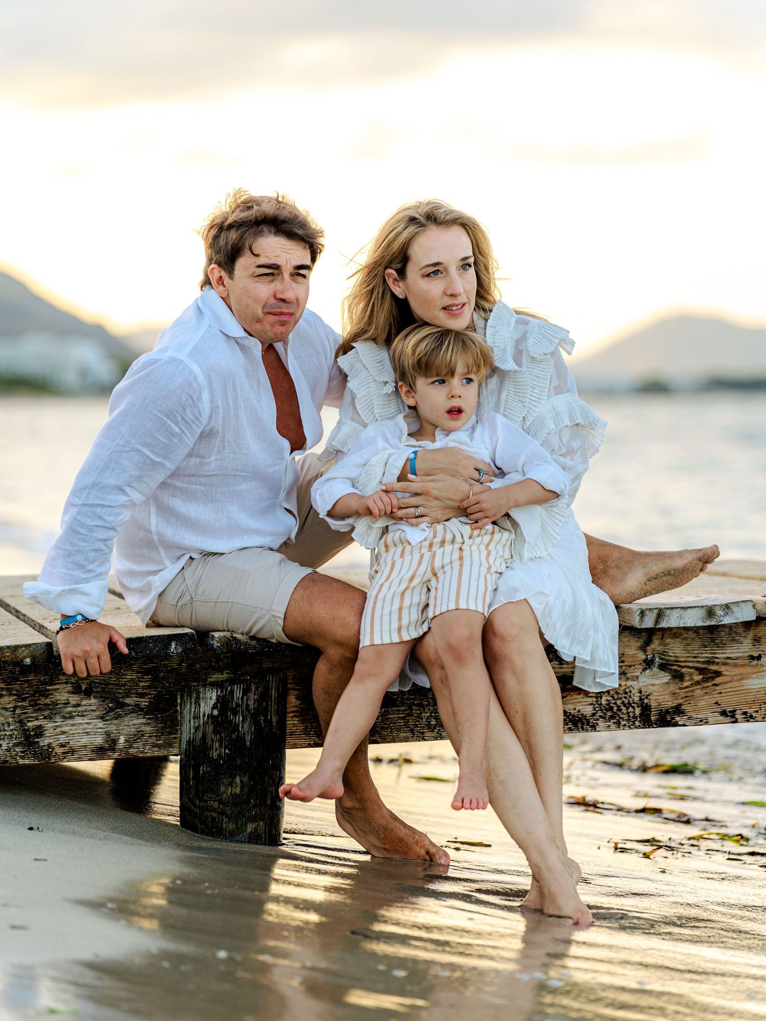 Happy family of three posing on a rustic beach dock at golden hour in coordinated white outfits.