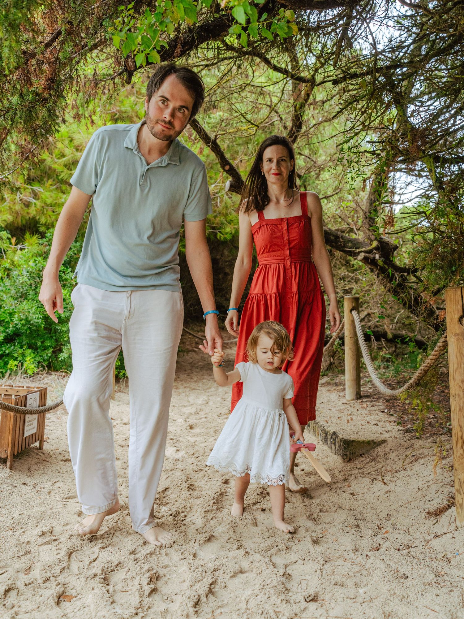 Young family with toddler daughter walking barefoot on sandy trail surrounded by green trees.