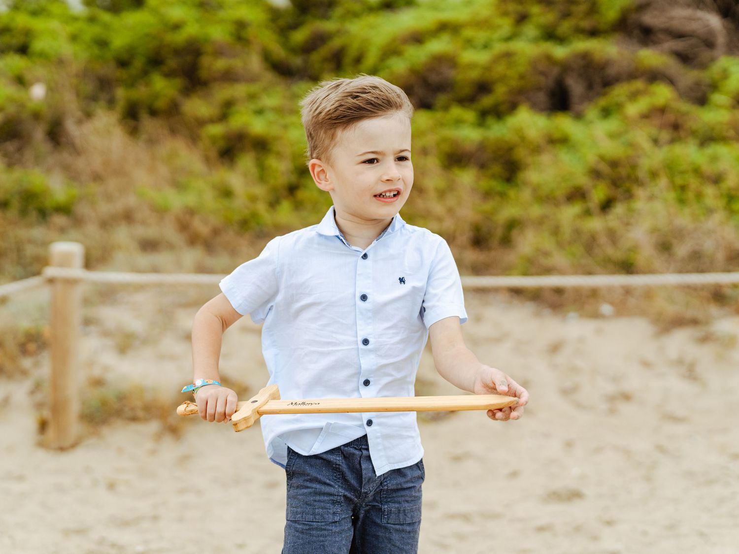 Young boy in light blue shirt confidently holding wooden stick at sandy beach fence.