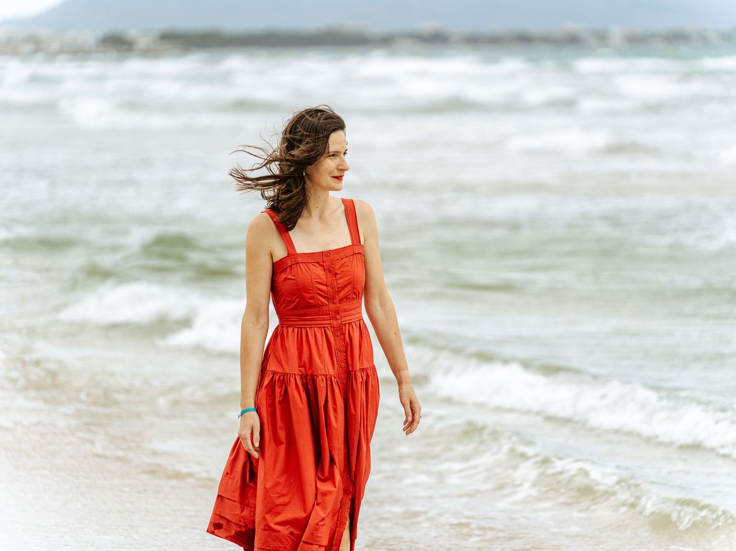 Smiling woman in a red dress walking along a breezy beach shoreline.