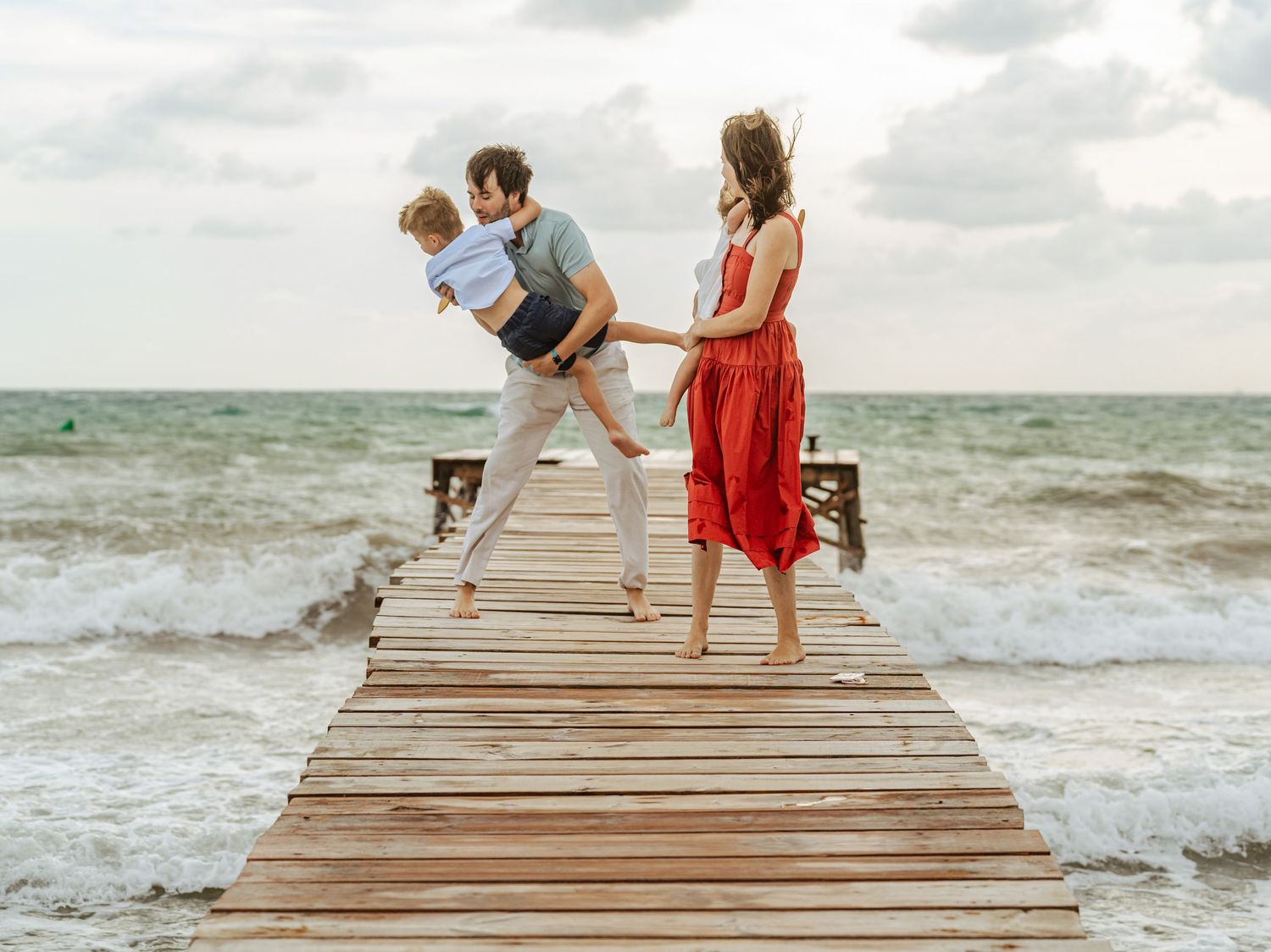 Family on beach pier, dad playfully leaning with son, mom in red dress, ocean backdrop.