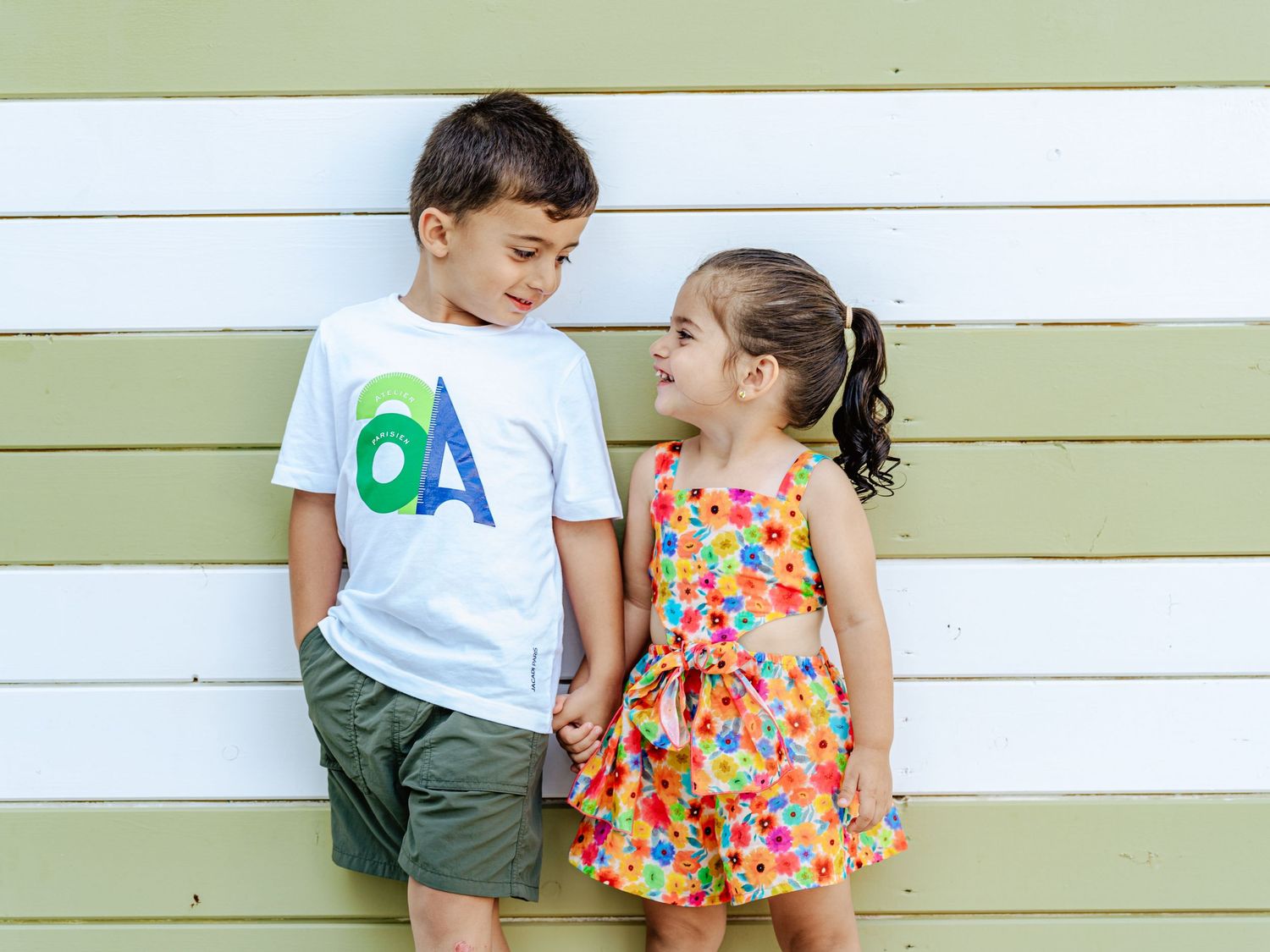 Young boy and girl holding hands and smiling at each other against a striped wooden wall.