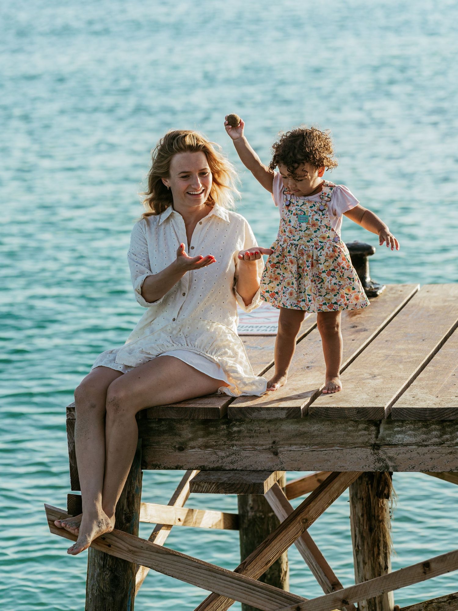 A woman in a white dress sits on a wooden pier while a young girl in a floral dress dances beside her over turquoise water.