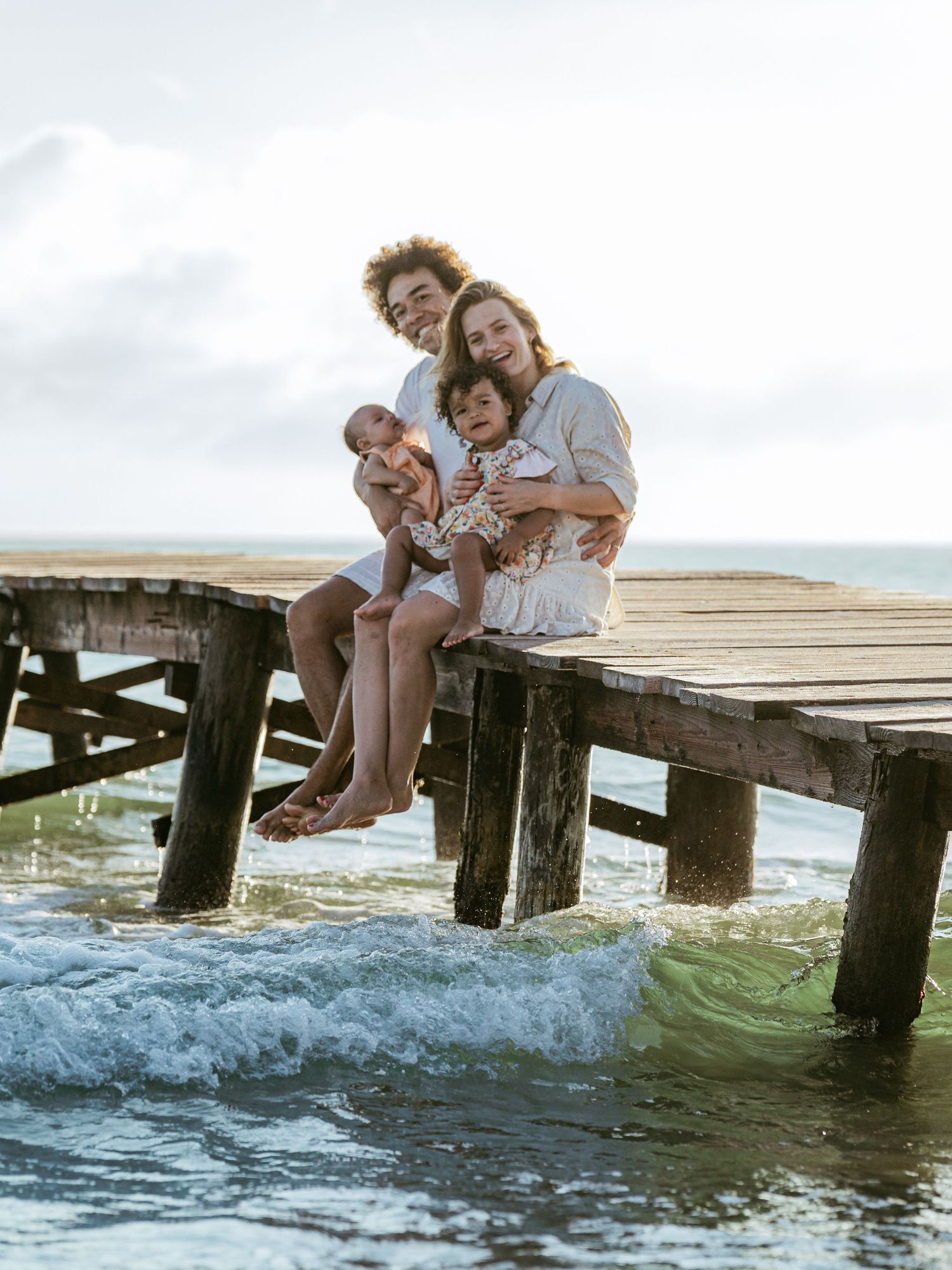 Family of four sitting on a wooden pier over ocean waves, parents holding baby and toddler, smiling at camera.