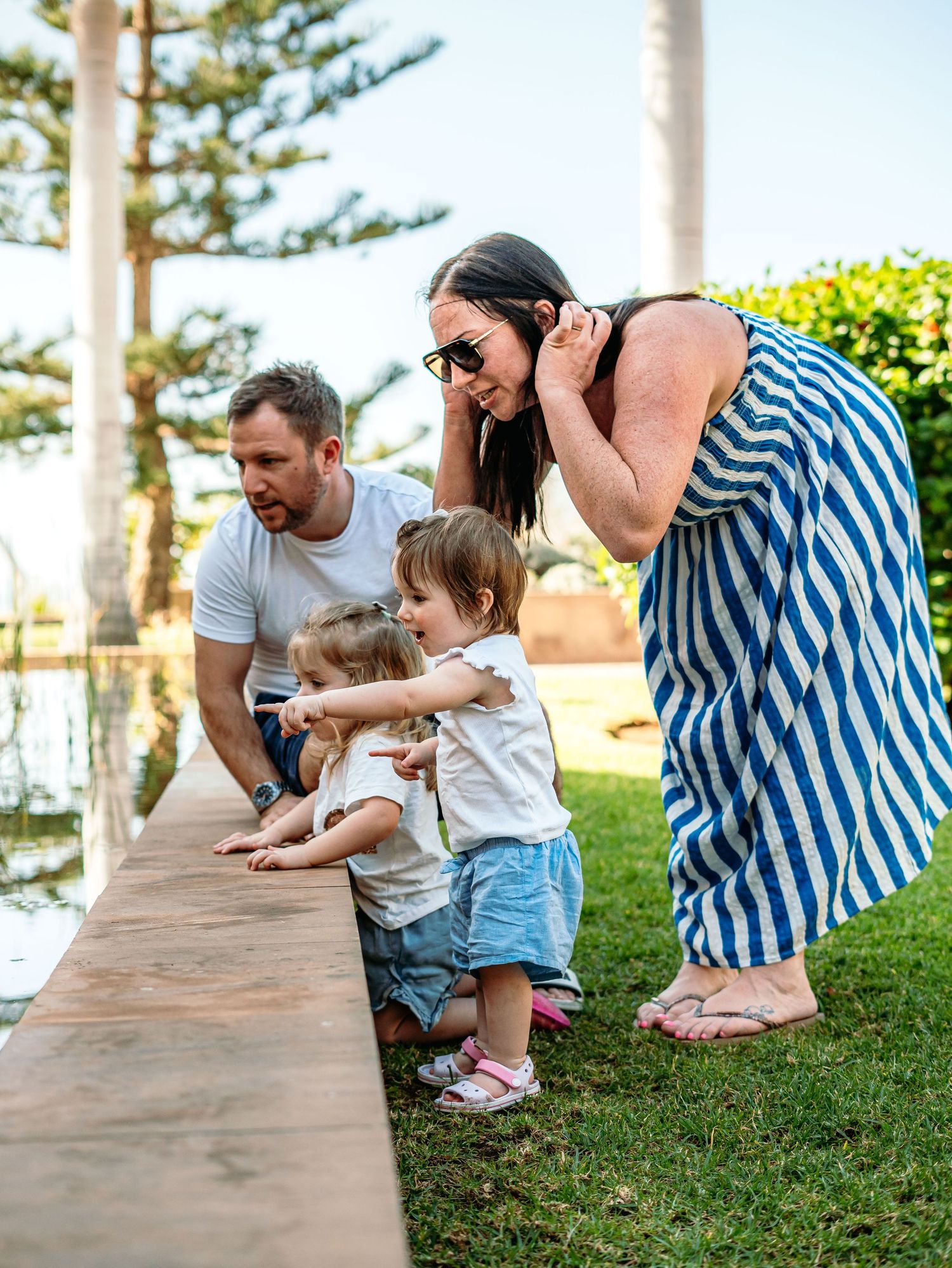 Family with two young children exploring a pond edge in a lush green garden with palm trees.