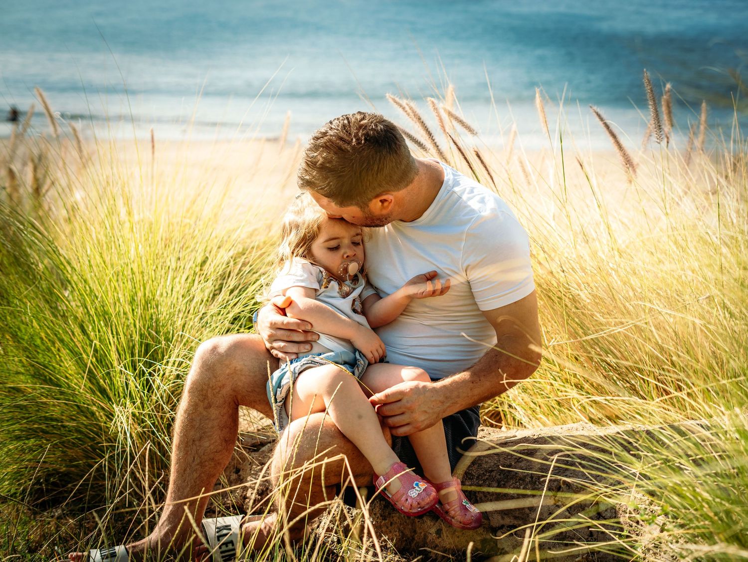 Loving father kissing young daughter while sitting in golden beach grass by ocean.