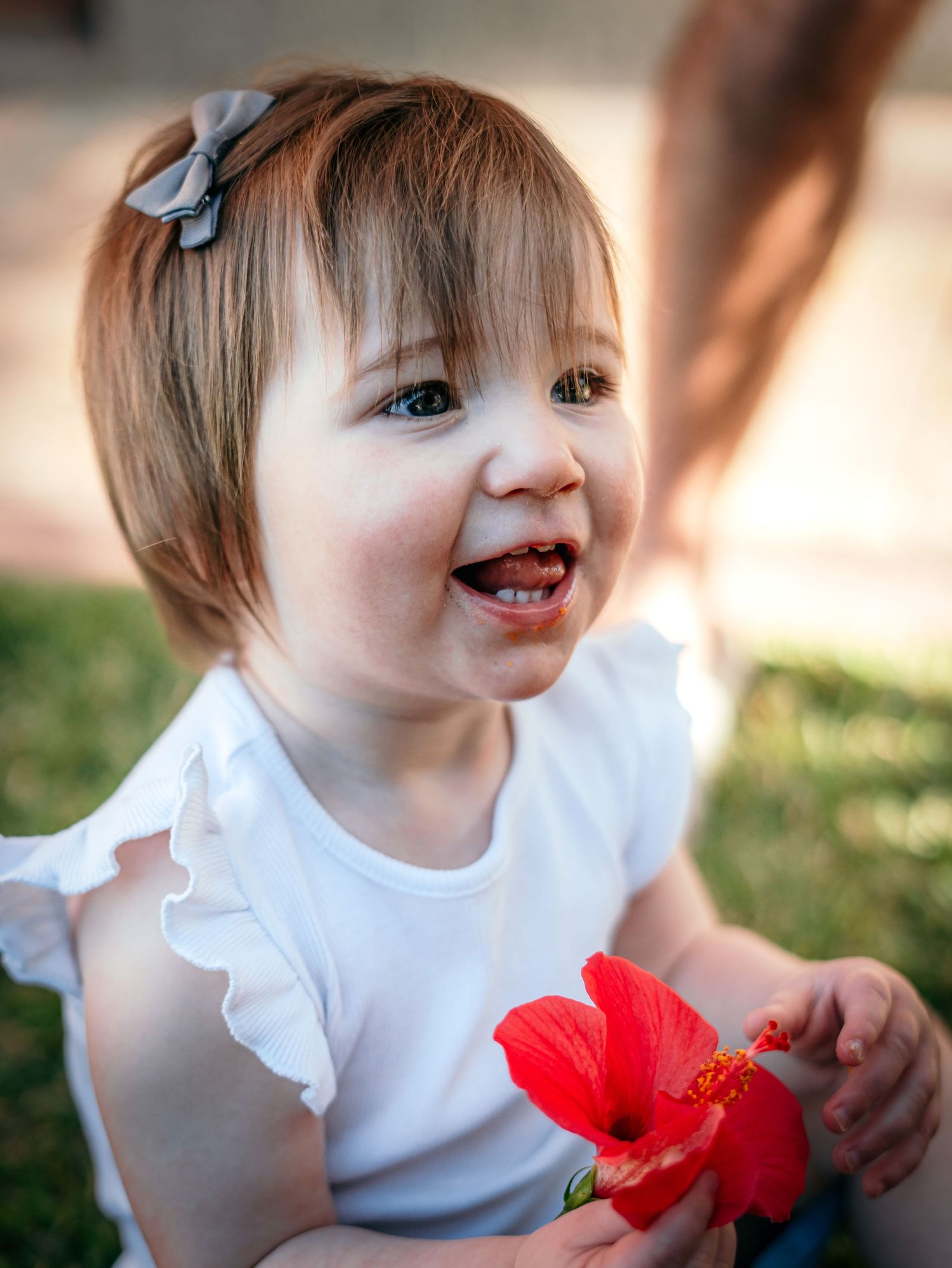 Happy baby girl with bow clip holds a red hibiscus flower while smiling outdoors.