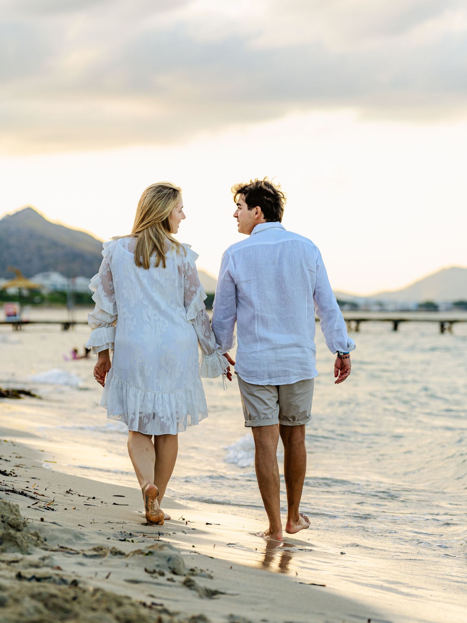 Couple in white outfits walking hand in hand along a beach with mountain views at sunset.
