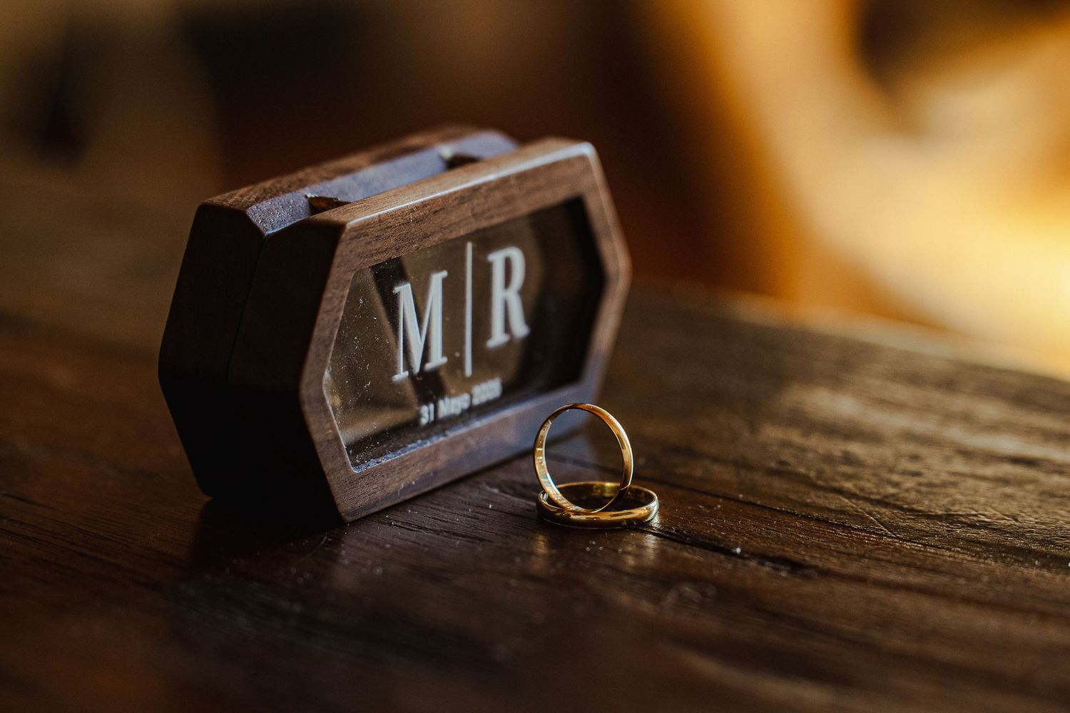 Gold wedding rings beside a personalized wooden ring box with monogram initials on a rustic wooden surface.