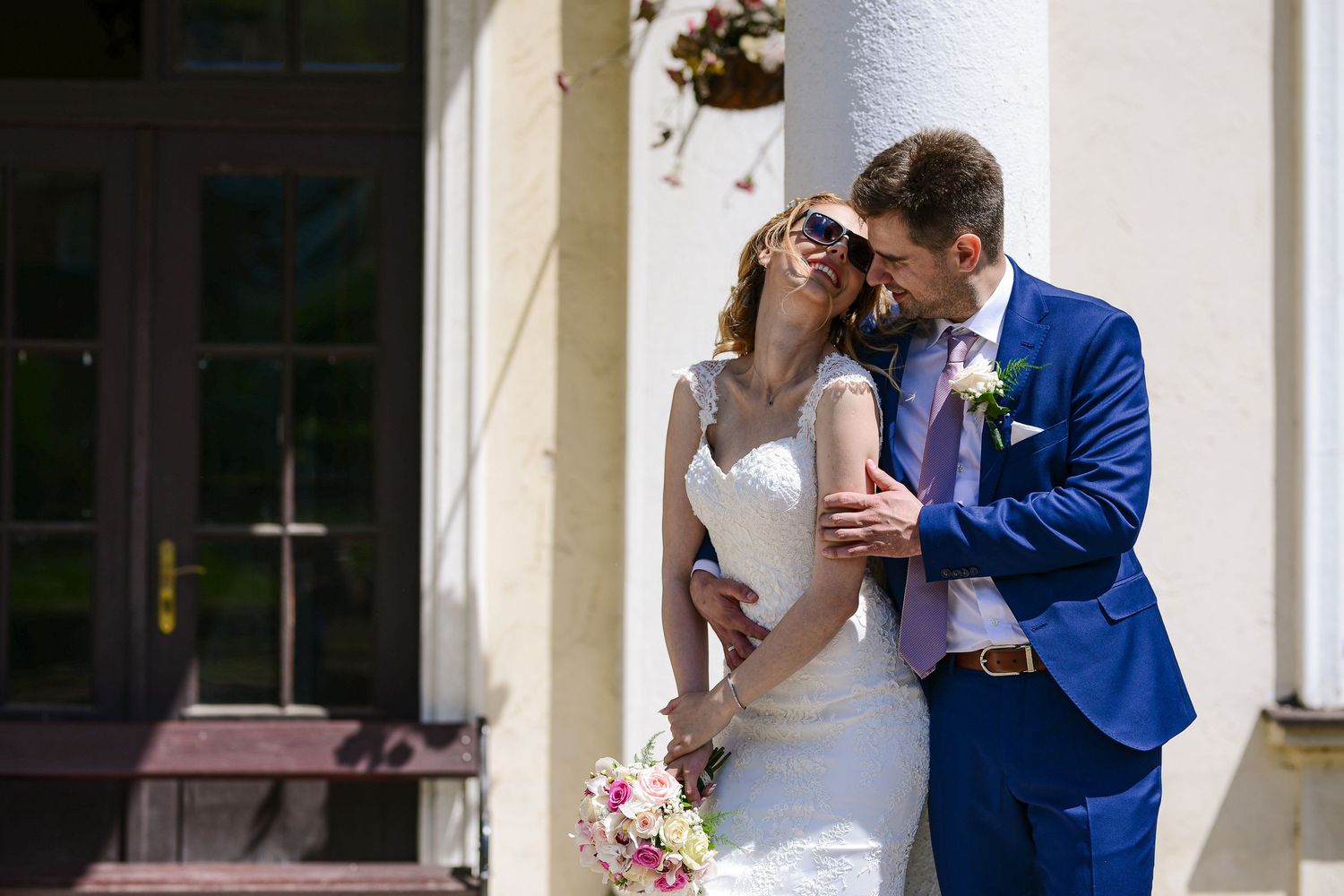 Bride in sunglasses and groom in blue suit share a tender moment beside white columns on a sunny wedding day.