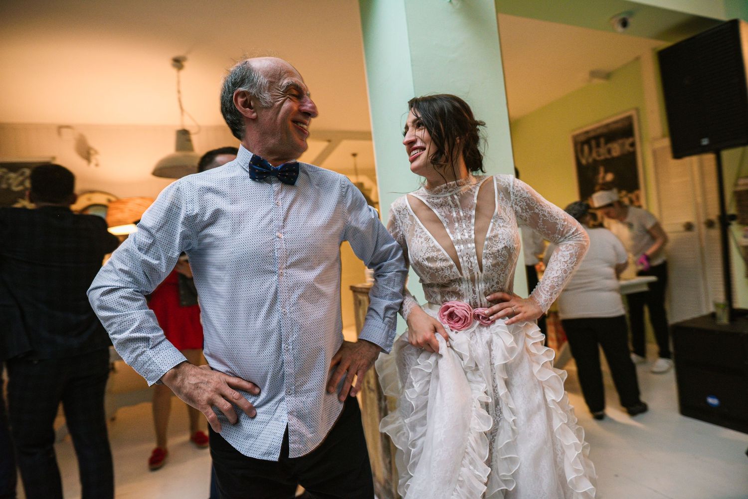 Elderly man in bow tie laughs with bride in lace wedding dress at reception.