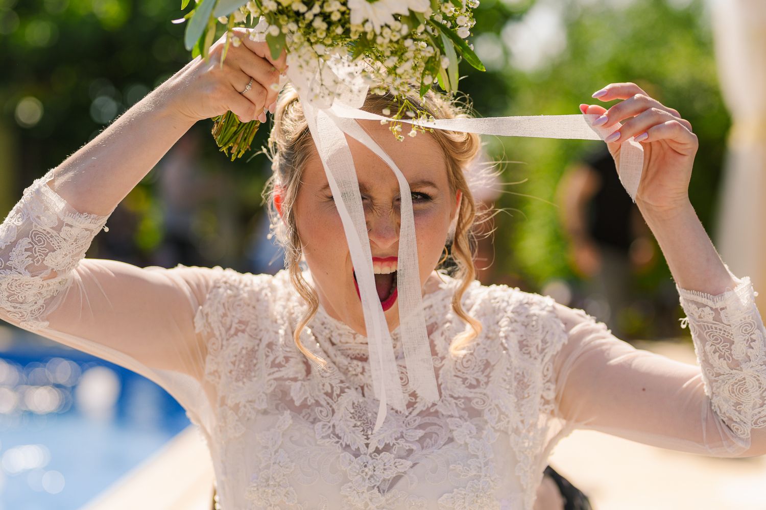 Bride in lace dress playfully holding white flower bouquet above her head outdoors.