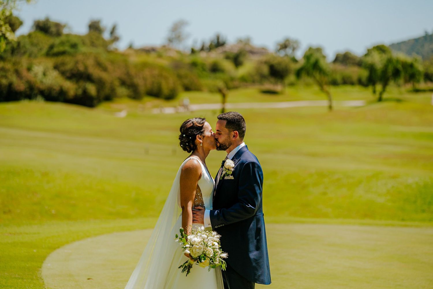 Newlyweds share a romantic kiss on lush green golf course under bright blue sky.