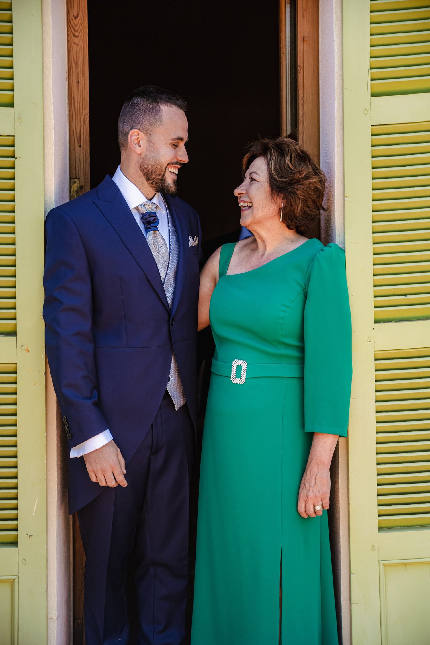 Groom in navy suit smiling with a woman in an elegant green dress in a doorway.