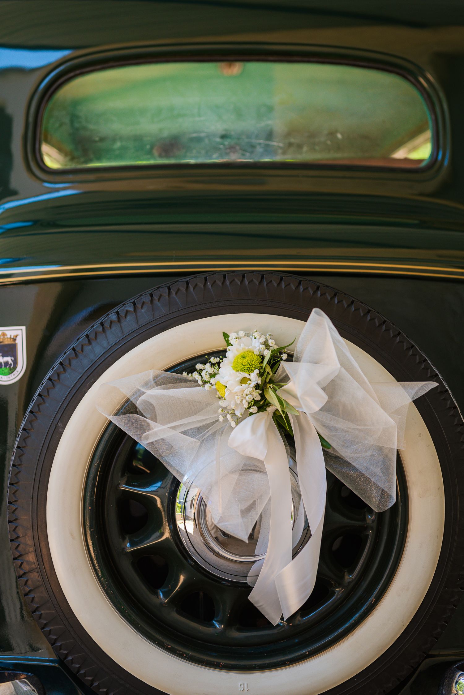 Vintage black wedding car decorated with white tulle bow and fresh flowers on wheel.