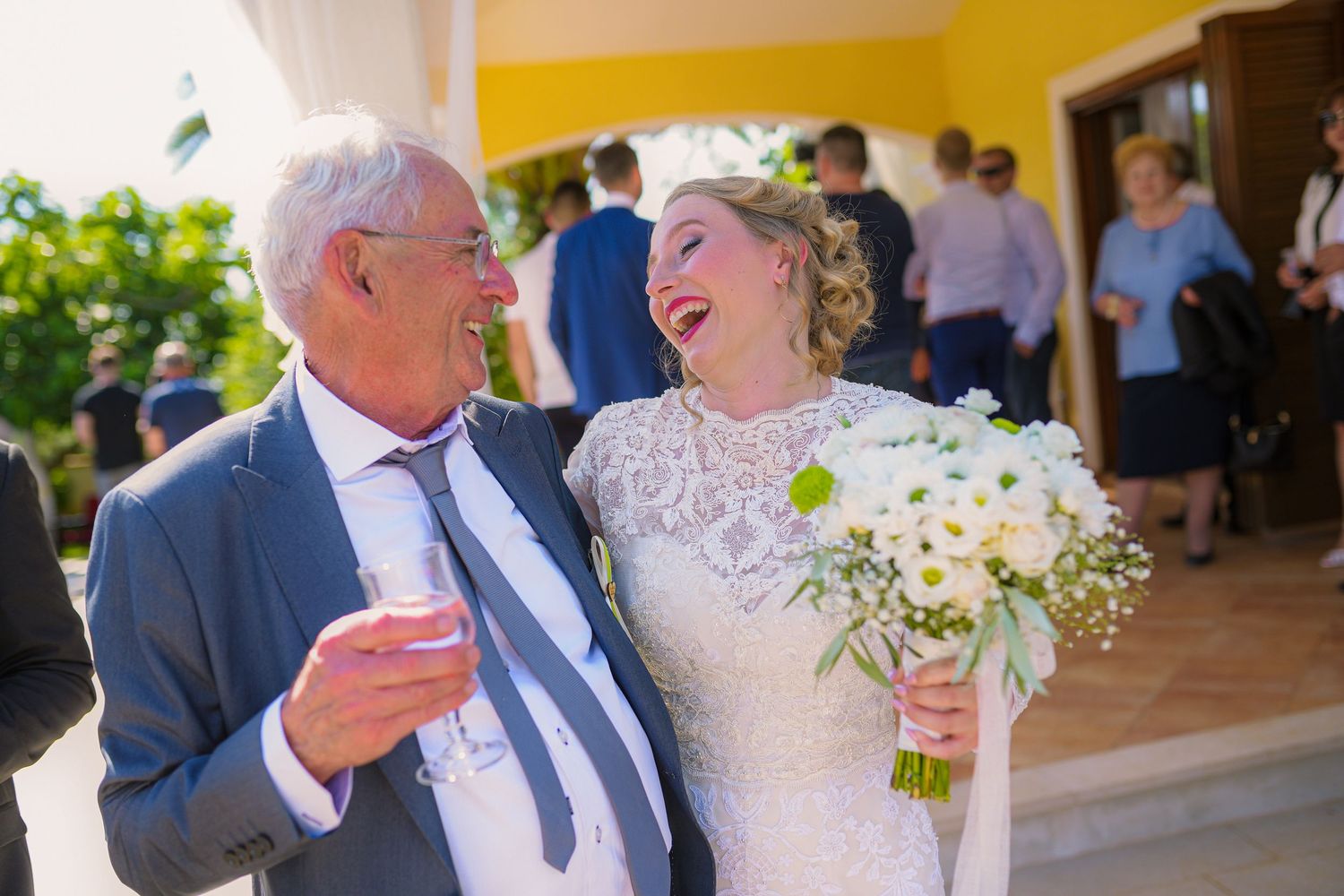 Bride laughing joyfully with older man in gray suit holding champagne at outdoor wedding reception.