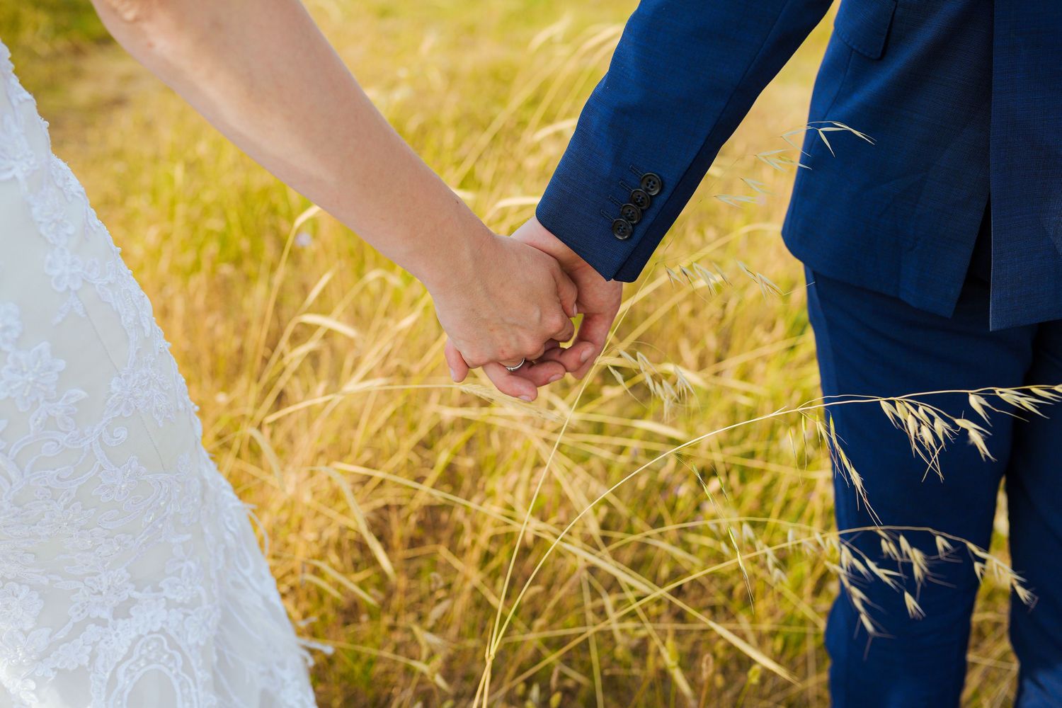 Close-up of bride and groom holding hands in a golden sunlit wheat field on their wedding day.
