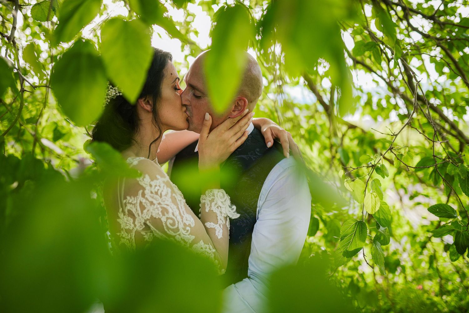 Bride and groom share a romantic kiss surrounded by lush green leaves in a garden setting.