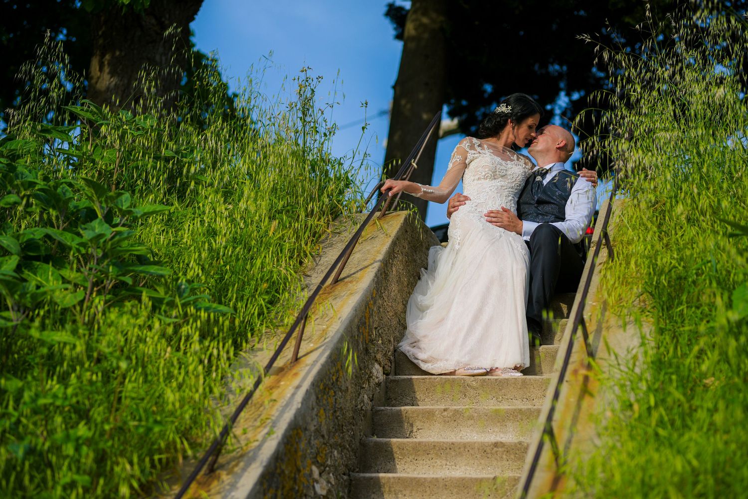 Bride and groom share a tender kiss on stone steps surrounded by tall green grass.