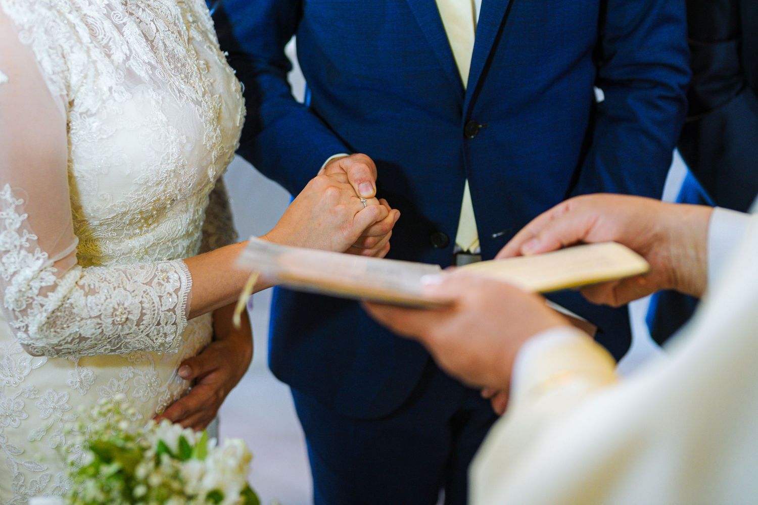 Groom in blue suit holding bride's hand during ring exchange at wedding ceremony.