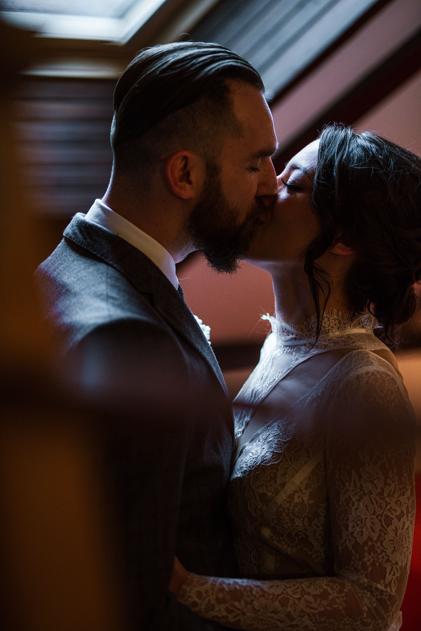 Bearded groom kissing his bride in a lace wedding dress in a beautifully lit intimate setting.