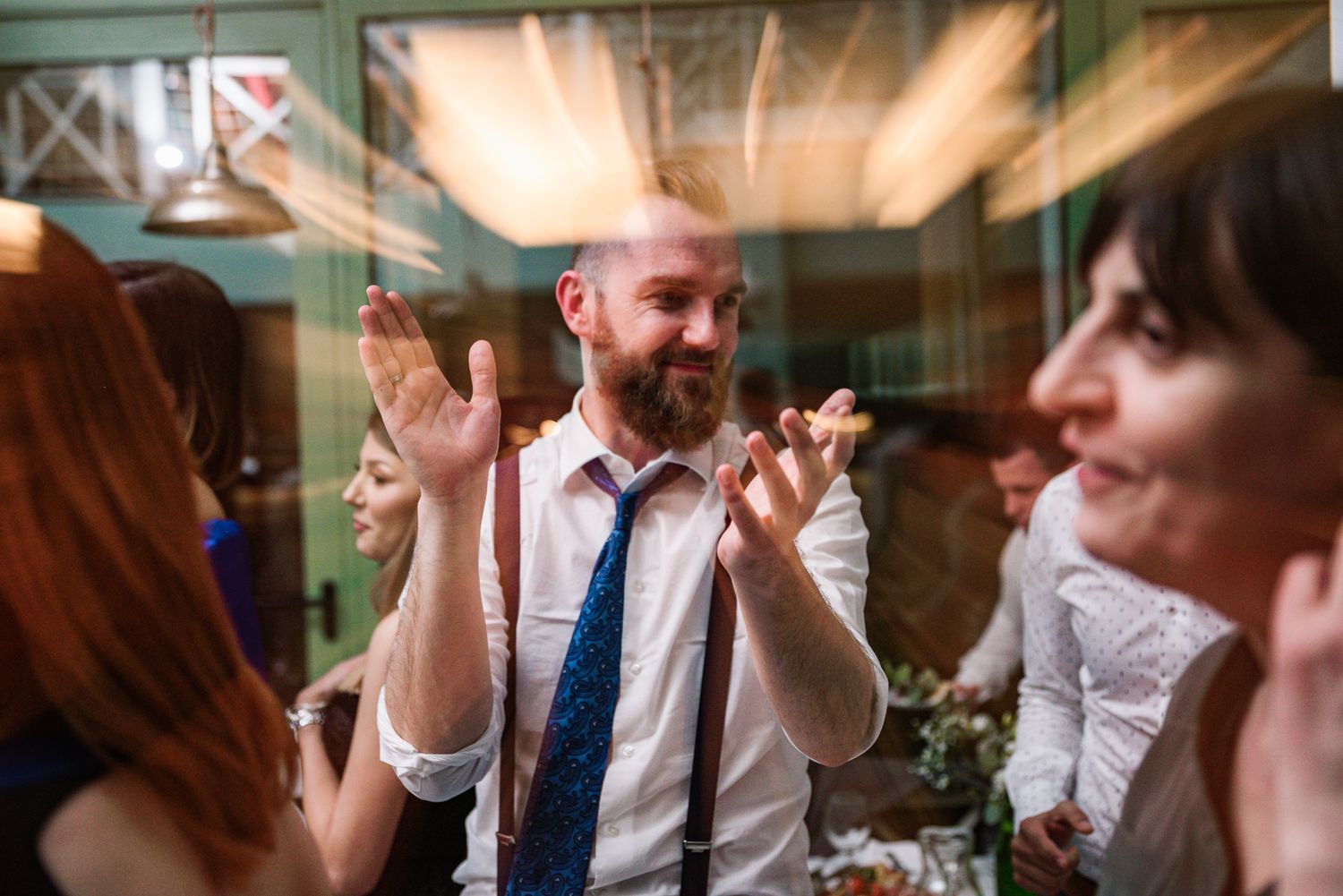 Bearded groom in suspenders and blue tie claps and celebrates at wedding reception.