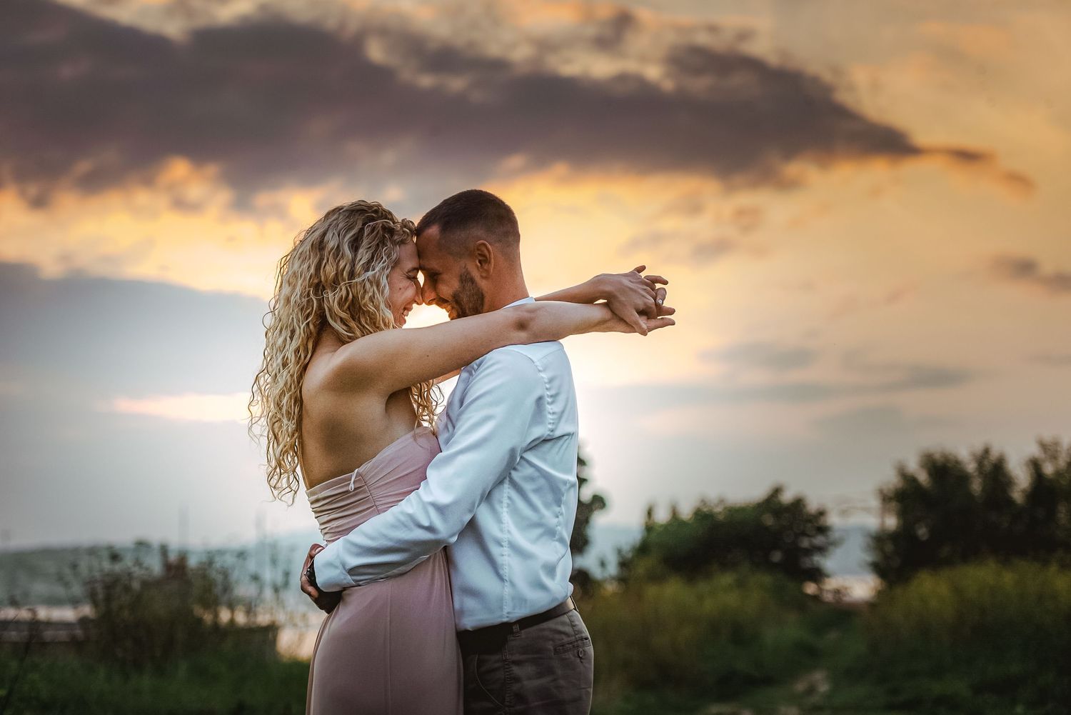 Couple dancing romantically at sunset with dramatic cloudy sky and scenic landscape in the background.
