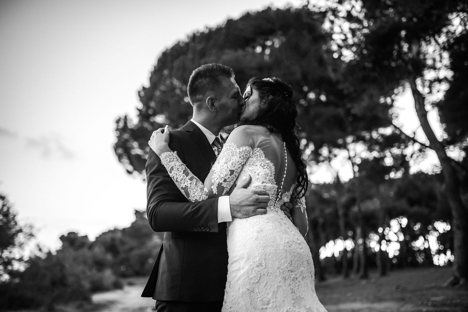 Black and white romantic wedding kiss between bride in lace gown and groom among tall pine trees.
