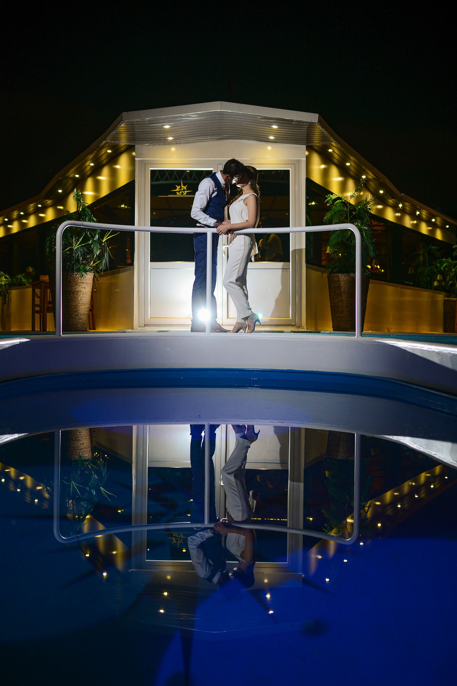 Wedding couple kissing on a lit bridge reflected in a pool during a romantic nighttime celebration.