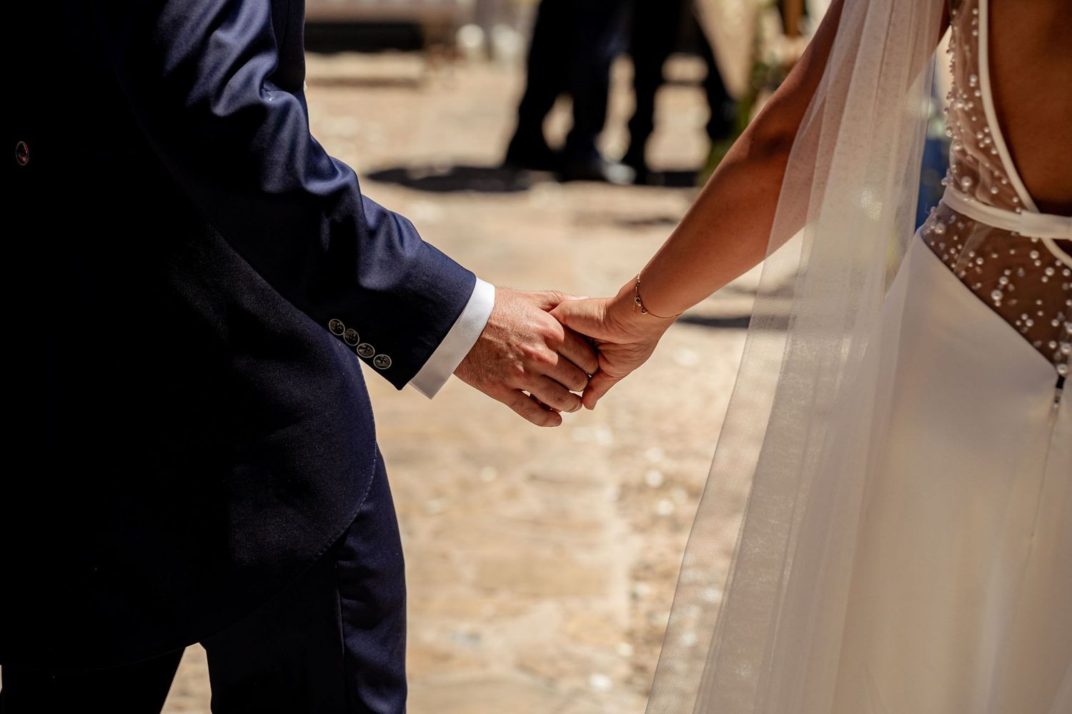 Bride and groom hold hands in sunlight after wedding ceremony, bride wearing flowing white gown.
