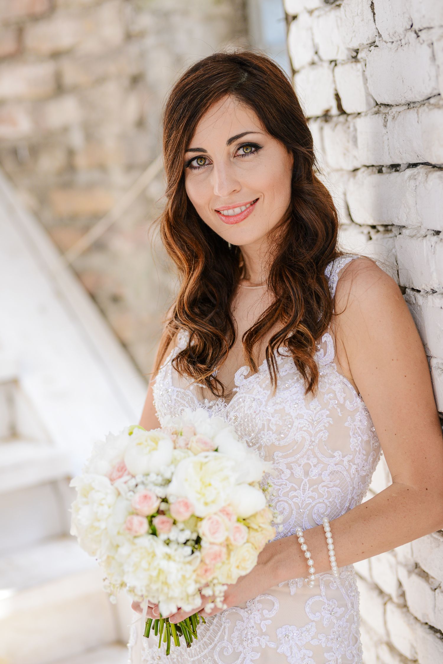 Beautiful bride in lace wedding dress holds a bouquet of white and pink flowers against a brick wall.