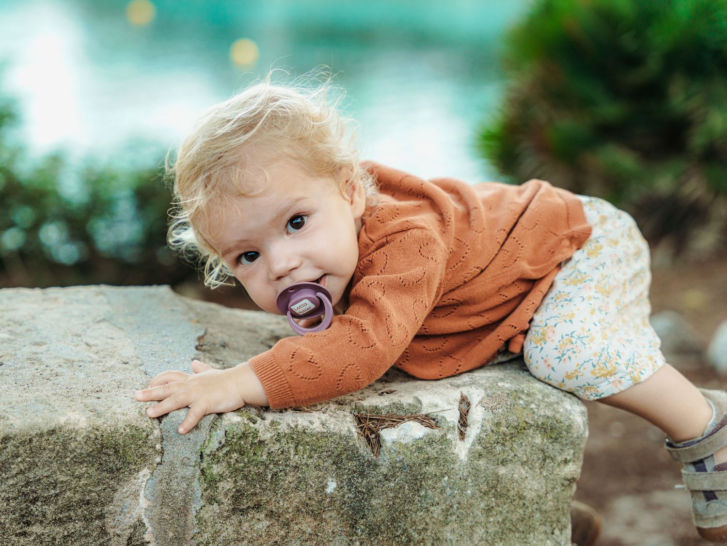 Curious toddler in orange cardigan climbing on a stone wall with a pacifier near turquoise water.