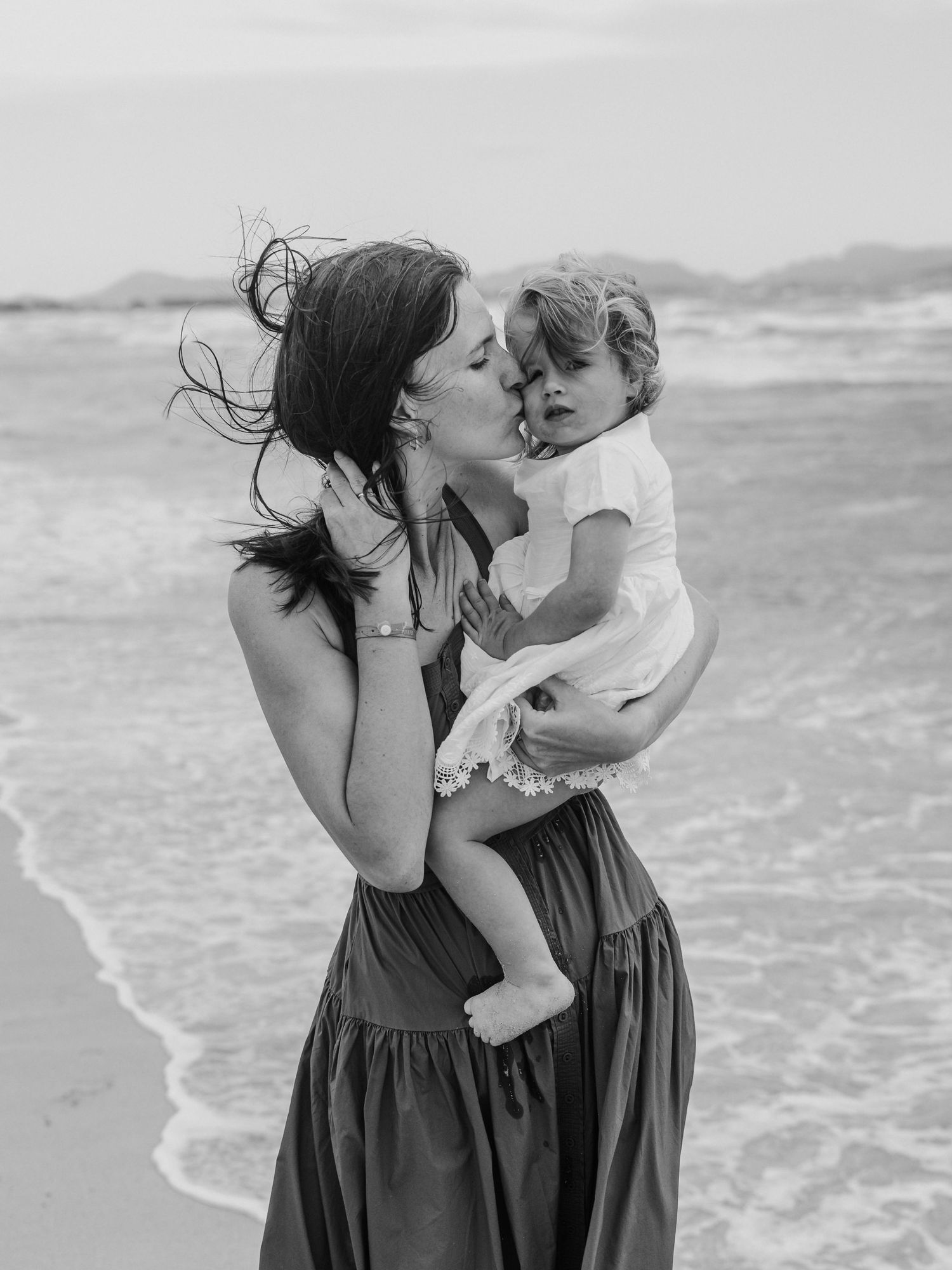 Black and white photo of mother kissing toddler on beach.