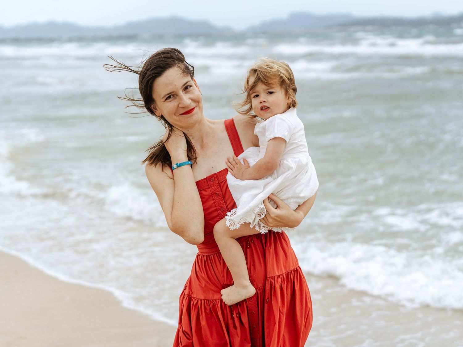 Mother in red dress carrying baby by ocean waves.