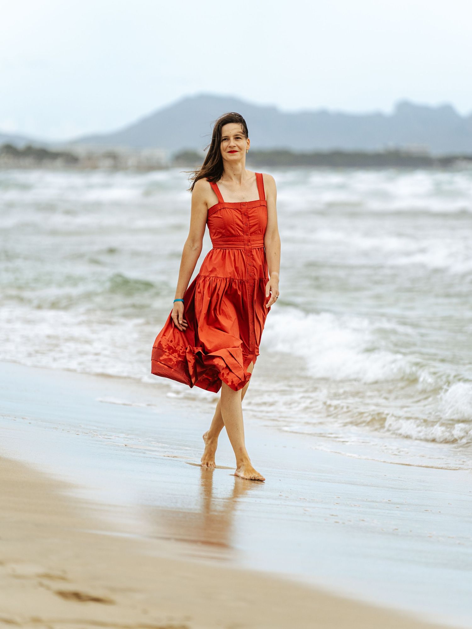 Woman in a red dress walking barefoot on a beach with mountains behind.