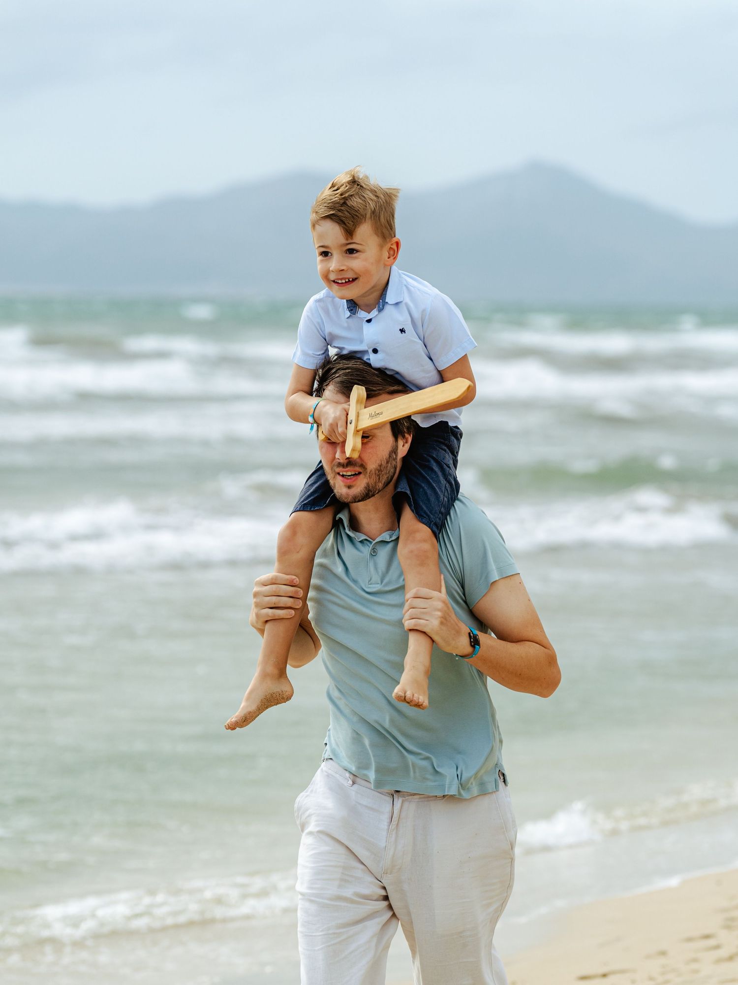 Smiling boy sits on fathers shoulders while enjoying a day at the beach with mountains behind.