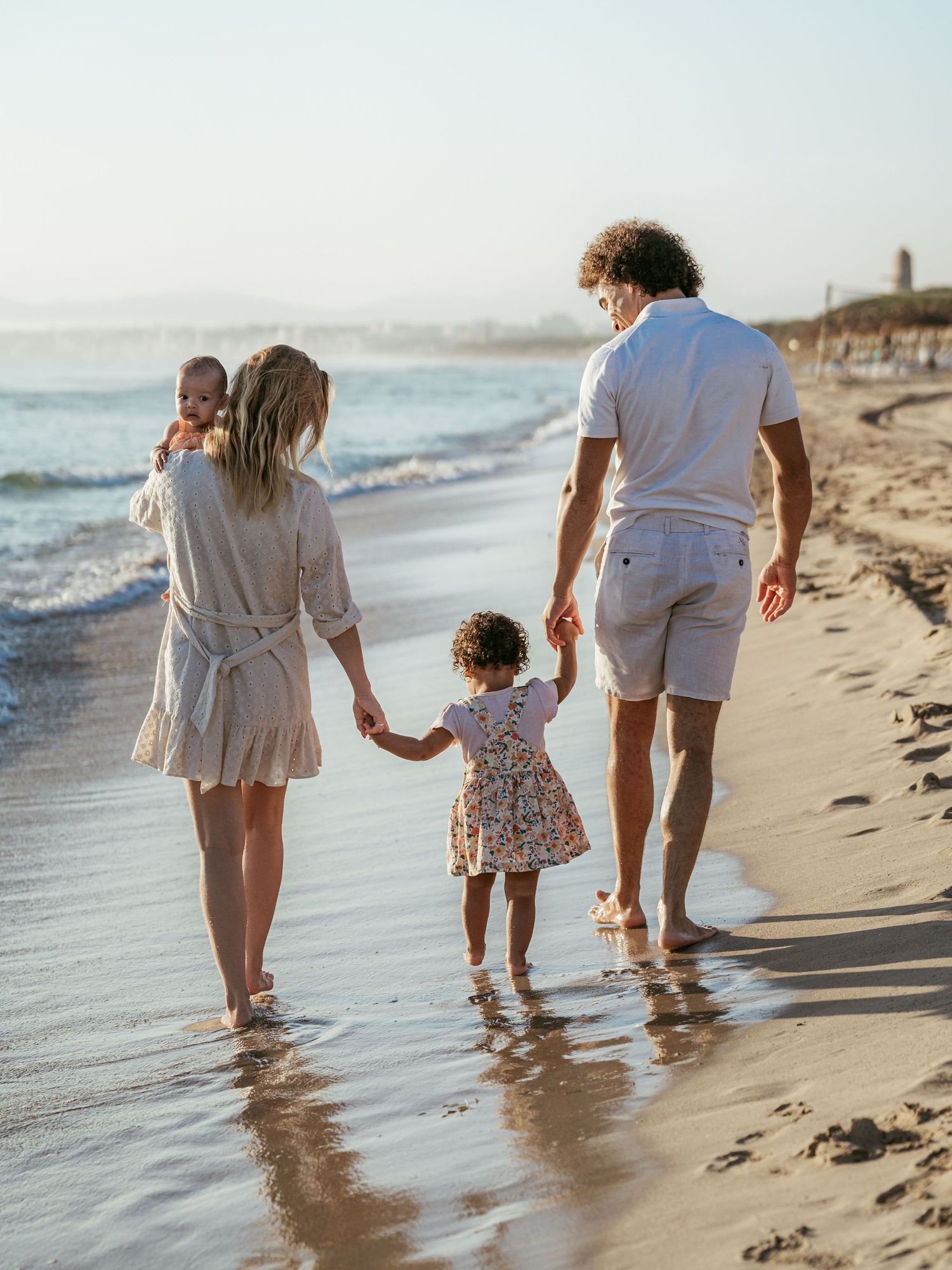 Family of four walking along a sunny beach, parents holding toddler's hands near the waves.