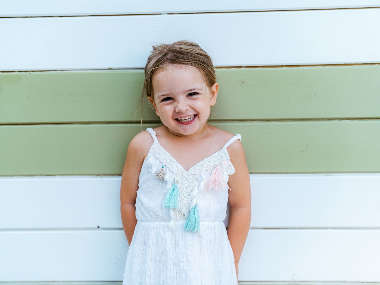 Happy young girl in white tassel sundress laughs joyfully while standing against a green and white wall.