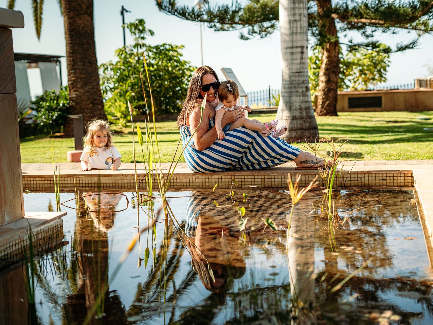 Mother and two young children sitting by a reflective garden pond surrounded by palm trees.