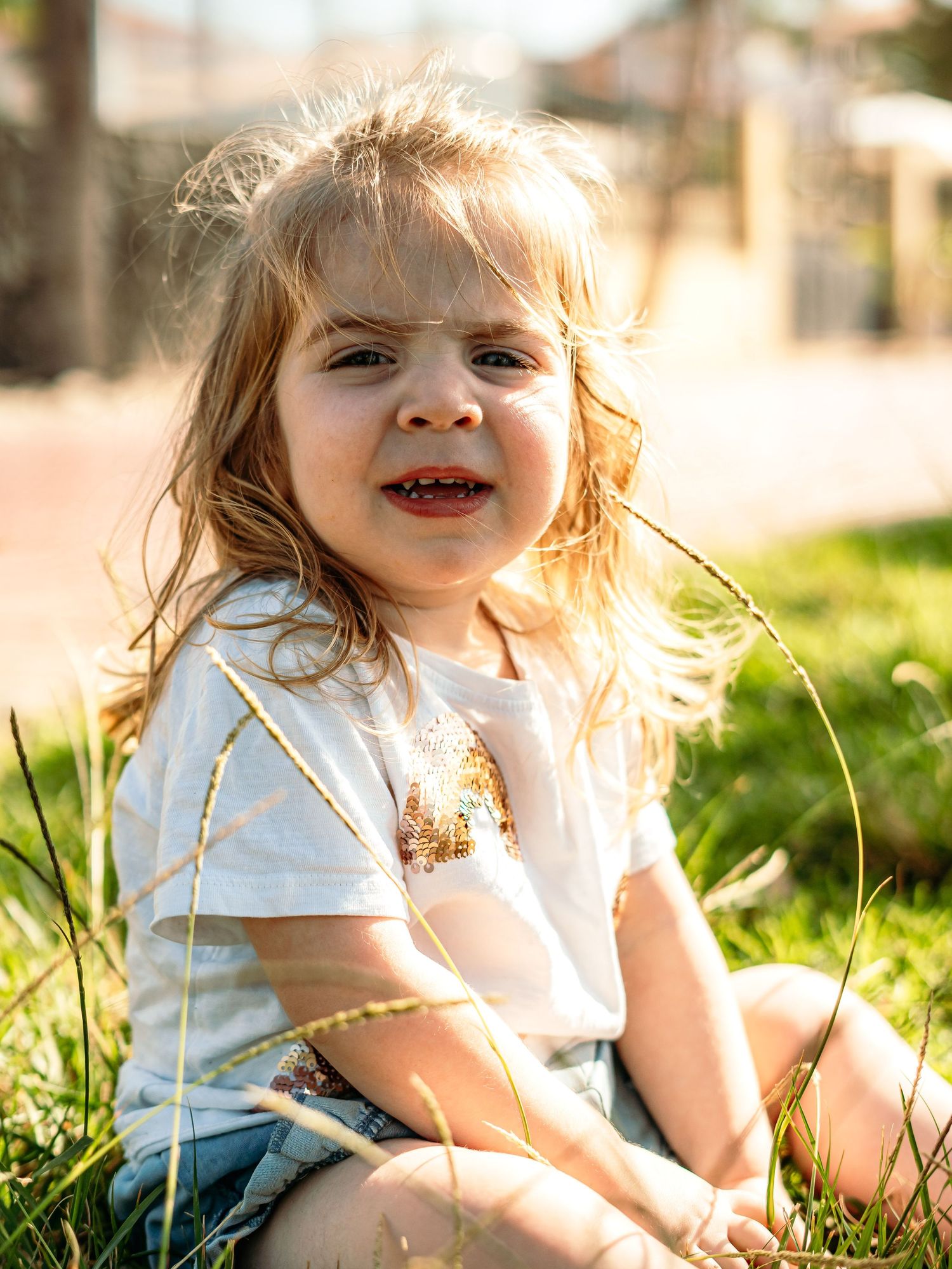 Cheerful blonde toddler girl sitting in sunlit grass smiling in white t-shirt and denim shorts.