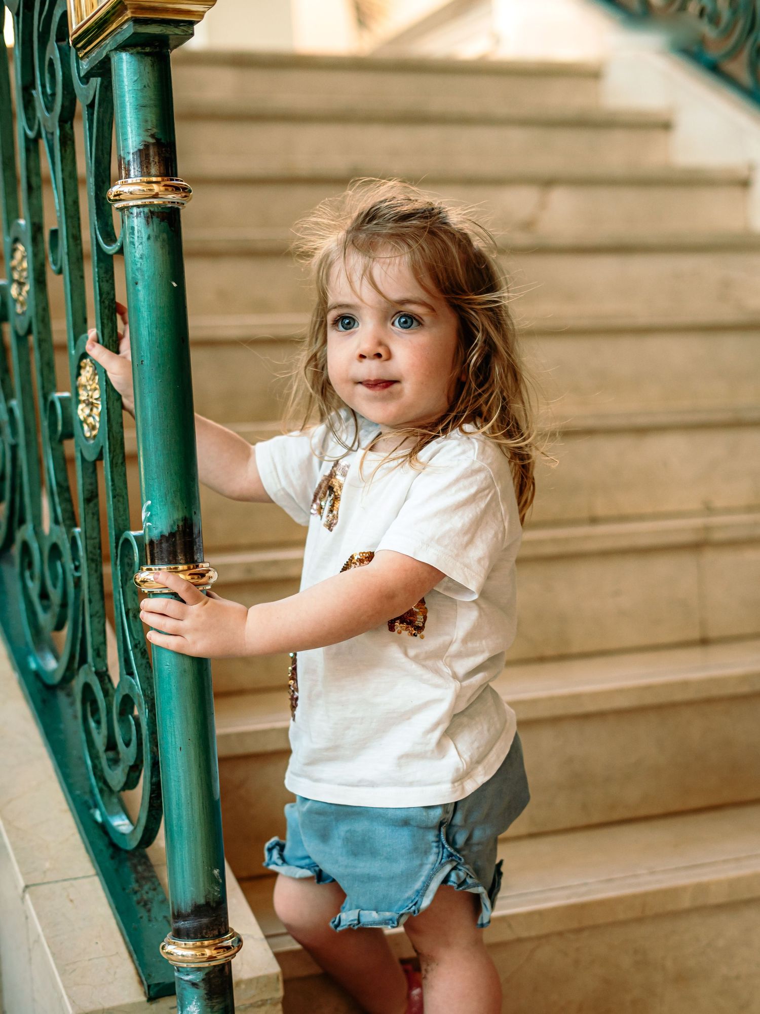 A young girl in a white tee and denim shorts holds a decorative green stair railing and looks at camera.