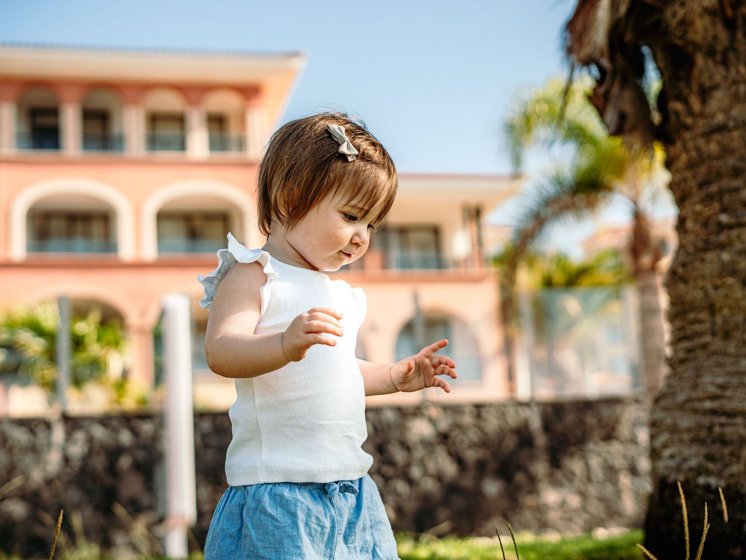 Cute toddler girl with hair clip in white sleeveless top and blue shorts standing outdoors by a pink resort.