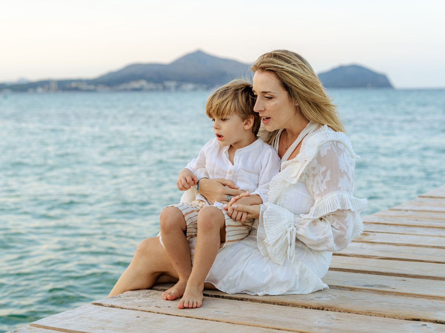 Mother and young son sitting quietly together on a sunlit wooden dock with mountain backdrop.