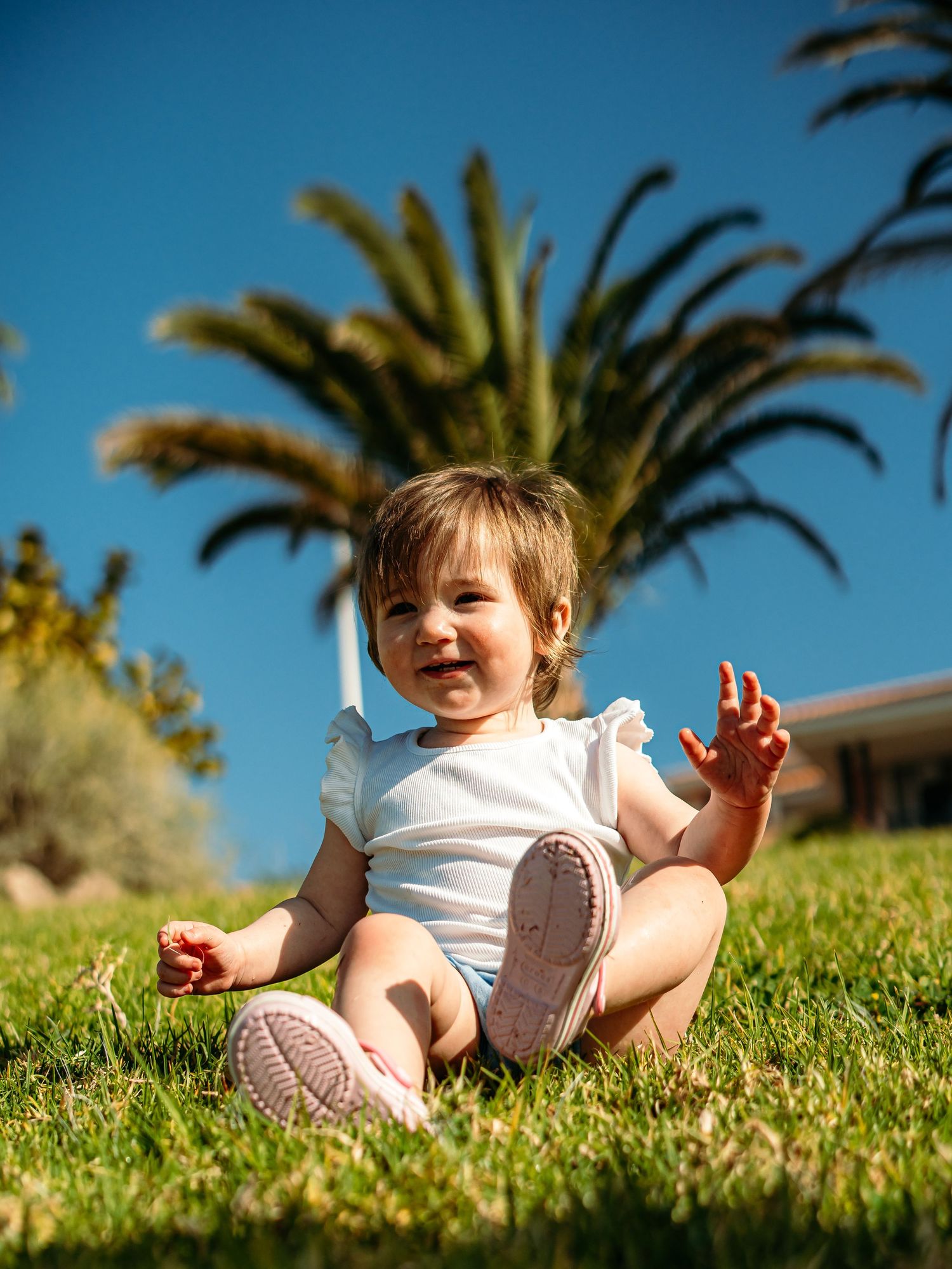 Happy baby girl sits on green grass smiling and waving with a tall palm tree and blue sky behind her.