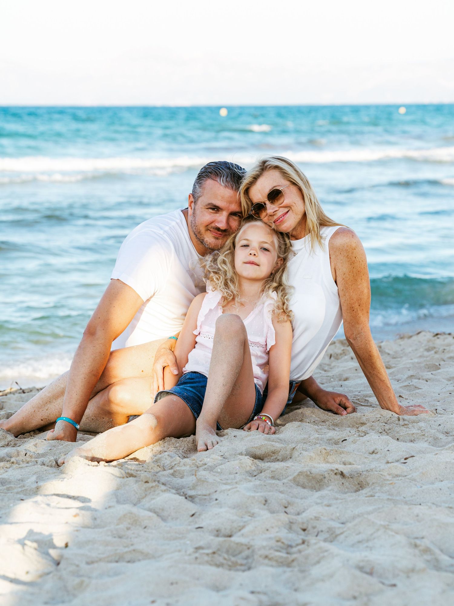 Happy family of three sitting together on sandy beach, smiling at camera with blue ocean in the background.