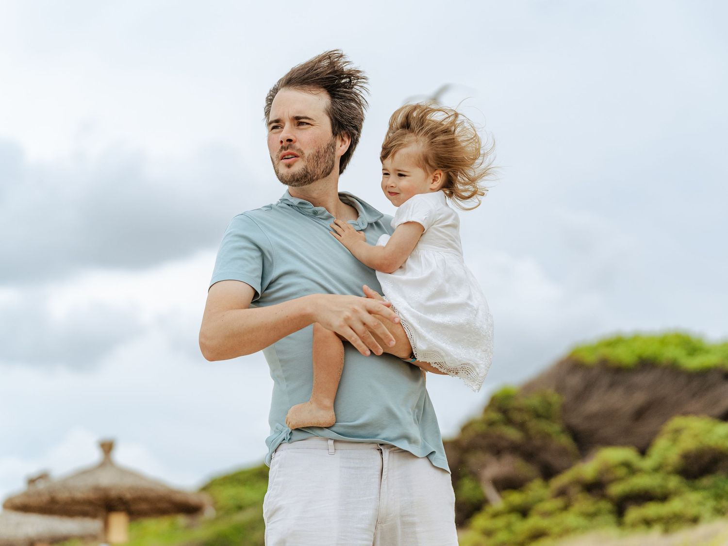 Father holding young child in arms on windy beach with cloudy sky background.