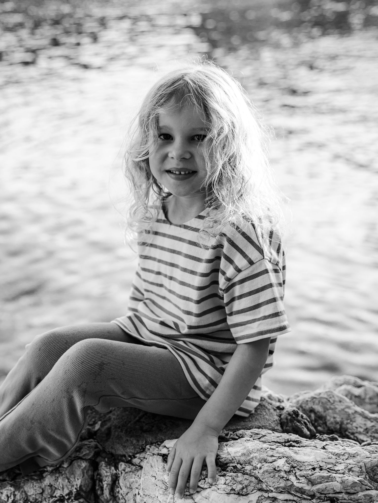 Black and white portrait of a curly-haired girl in striped shirt sitting on rocks by water.